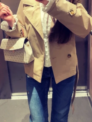  beige trenchcoat med bälte - Snygg kort beige trenchcoat med dubbelknäppning och bälte i midjan. Klassisk krage och stora knappar i kontrastfärg. Jackan har en stilren och tidlös look, perfekt att matcha med jeans eller kostymbyxor. Tillverkad i ett slätt tyg som ger en elegant känsla.