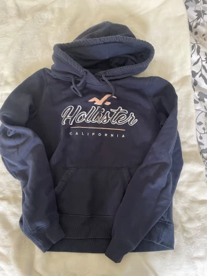 Mörkblå hoodie från Hollister - Mörkblå hoodie från Hollister med broderad logga. Klassisk känguruficka framtill och justerbar huva med dragsko. Mjuk insida och ribbade muddar vid ärmar och nederkant. Perfekt för en avslappnad och stilren look. 