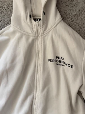 Vit hoodie Peak Performance - Säljer en vit hoodie från Peak Performance med dragkedja framtill och broderad logga på bröstet. Hoodien har huva med snörning och två fickor fram. Tillverkad i mjukt bomullsmaterial och har ribbade muddar vid ärmslut och nederkant. Den är i storlek L men skulle säga att den är väldigt liten i storleken, så mer en S 