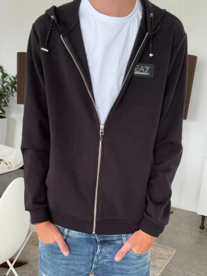 Svart zip hoodie från EA7 Emporio Armani - Snygg svart hoodie med dragkedja från EA7 Emporio Armani. Hoodien har huva med dragsko, ribbade muddar och en liten logopatch på bröstet. Tillverkad i mjukt material som passar perfekt till jeans eller joggers. Passar större S