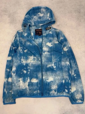 Blåvit vindjacka med huva Woolrich - Snygg blåvit vindjacka från Woolrich med unikt mönster som påminner om moln. Jackan har hel dragkedja framtill, huva och två sidofickor. Perfekt för dig som vill sticka ut med en cool och modern look. Passar M-L. Ställ gärna frågor!🙂