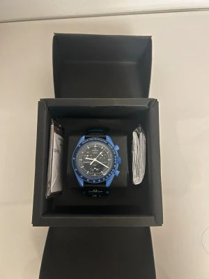 Omega x Swatch blå kronografklocka - Säljer en snygg Omega x Swatch kronografklocka med blå boett och svart urtavla. Klockan har vita visare och detaljer samt svart band med Omega och Swatch-logga. Perfekt för dig som gillar sportig och modern stil. Vid snabb köp kan priset minskas.