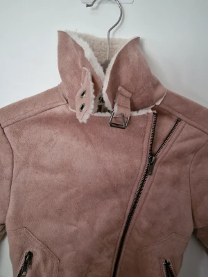 Rosa teddyjacka från H&M storlek 135/68 - Säljer en snygg rosa teddyjacka från H&M med mjukt, vitt foder i teddy och coola detaljer som sned dragkedja, höga krage med spänne och fickor med dragkedja. Perfekt för dig som vill ha en trendig och mysig jacka till hösten eller vintern.