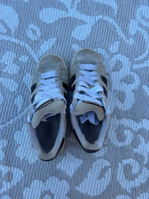 Adidas Campus beige sneakers - Snygga Adidas Campus sneakers i beige mocka med klassiska svarta ränder på sidorna och vit sula. Skorna har vita skosnören och gummisula för en retro look. Perfekta för dig som gillar streetwear och tidlös stil.