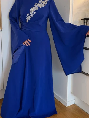 Blå långklänning med vida ärmar - Elegant blå långklänning med vida, svepande ärmar och dekorativt broderi i vitt över bröstet. Klänningen har en markerad midja och en stilren, flytande siluett som ger ett lyxigt intryck. OBS! Modellen är xs och bär storlek L