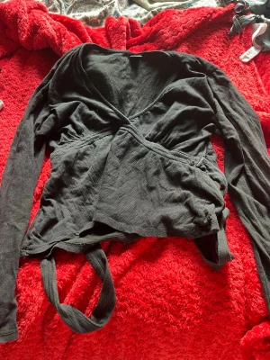 Svart omlottopp med långa ärmar - Snygg svart omlottopp med långa ärmar och knytband i midjan. Toppen har en djup v-ringning och är gjord i ett mjukt, tunt material som känns skönt mot huden. Perfekt för lager på lager och enkel att styla till olika outfits.