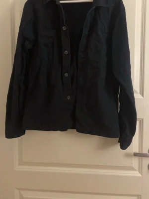 Svart långärmad skjorta med knappar - Stilren svart skjorta med långa ärmar och klassisk krage. Skjortan har knäppning framtill och två bröstfickor. Perfekt till både jeans och chinos för en clean look.