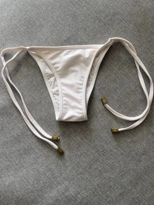Vit bikiniunderdel med knytband från Brasilien  - Snygg vit bikiniunderdel med smala knytband i sidorna och små detaljer på snörena. Klassisk modell från Brasilien som är enkel att matcha med olika överdelar. Perfekt för lata dagar på stranden eller vid poolen. #bikini #ciamaritima