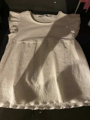 Vit topp med volangärm från 157 - Säljer en söt vit topp från 157 med korta volangärmar och rund hals. Tyget är strukturerat och ger en trendig look. Perfekt att matcha med jeans eller kjol för en fräsch outfit.