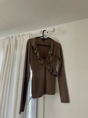 Brun volangtop från Amisu - Säljer en brun långärmad topp från Amisu med snygg volangdetalj längs den v-ringade halsen. Toppen har en slim passform och är tillverkad i ett mjukt, stretchigt material som sitter skönt på kroppen. Perfekt för dig som gillar stilrena men ändå unika plagg.