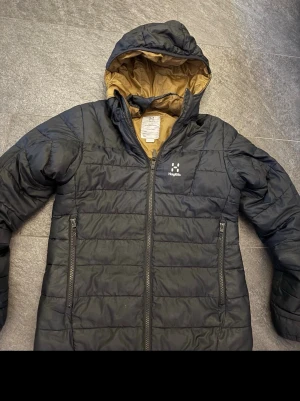 Svart dunjacka med huva från Haglöfs - Säljer en svart dunjacka från Haglöfs i storlek 164 jackan har en stilren look med avtagbar luva i riktigt bra skick perfekt för hösten 