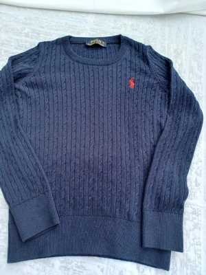 Mörkblå stickad tröja från Polo Ralph Lauren - Mörkblå stickad tröja från Polo Ralph Lauren med klassiskt kabelstickat mönster och röd broderad logotyp på bröstet. Rund halsringning och långa ärmar. Perfekt för dig som gillar stilrena och tidlösa plagg.