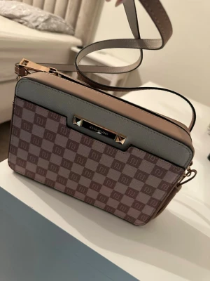 Rutigt axelremsväska från River Island - Snygg handväska från River Island med rutigt mönster i beige och brunt. Väskan har detaljer i ljusgrått och nude, guldfärgade metalldetaljer och en justerbar axelrem. Perfekt storlek för det viktigaste och stängs med dragkedja. Materialet är konstläder.