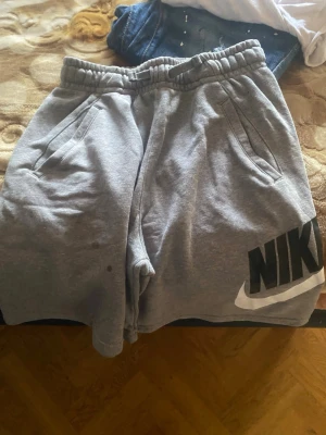 Gråa sweatshorts från Nike - Gråa sweatshorts från Nike med resår i midjan och snörning. Stora fickor på sidorna och en tydlig svart Nike-logga med vit swoosh på ena benet. Perfekta för chill eller träning, tillverkade i mjuk bomullsblandning.