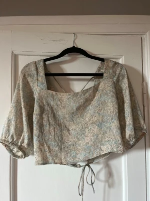 Blommig blus med puffärm och knyt - Supersöt blommig blus med ljusa toner, puffiga trekvartsärmar och knyt i midjan. Toppen har fyrkantig ringning och öppen rygg med tunna band som korsas. Perfekt för dig som gillar romantisk och trendig stil.