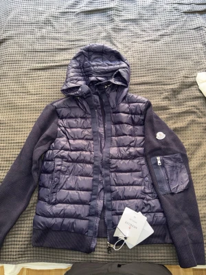 Moncler Cardigan med nfc  - Snygg mörkblå pufferjacka från Moncler med huva och stickade ärmar. Jackan har dragkedja framtill, fickor med knappar och en extra ficka med dragkedja på ärmen. Perfekt för kyliga dagar och ger en sportig look.