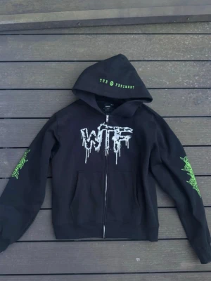Svart hoodie med tryck och dragkedja - Säljer en svart hoodie med dragkedja och huva. På bröstet står det 'WTF' i vitt, och ärmarna har gröna grafiska tryck. Huvan har texten 'The Promised' i grönt. Baksidan har stort tryck med turnéstäder och grön demonliknande grafik. Perfekt för dig som gillar streetwear.