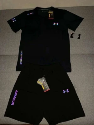 Svart träningsset från Under Armour - Snyggt svart träningsset från Under Armour med t-shirt och shorts. T-shirten har logga på bröstet och textdetaljer på axel och sida. Shortsen har dragsko i midjan, logga och texten 'ARMOUR' på benet. Perfekt för gymmet eller löprundan.