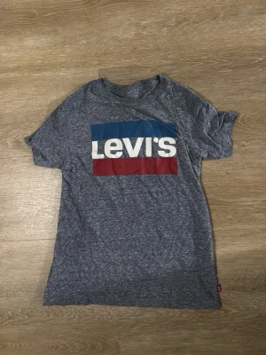Grå Levi's t-shirt med logga - Säljer en gråmelerad t-shirt från Levi's med stor färgglad logga i blått, vitt och rött på bröstet. Klassisk rund hals och korta ärmar. Perfekt för en avslappnad och stilren look.