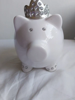 Vit spargris med silverkrona - Säljer en vit spargris i keramik med en glittrig silverfärgad krona och diamanter på kronan .Perfekt för att spara mynt och sedlar, och blir en snygg detalj i rummet. Myntinkast finns på ovansidan.