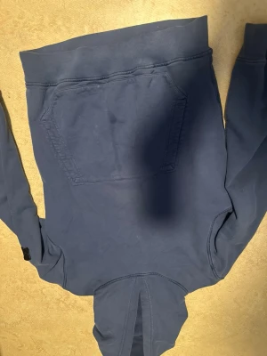Blå hoodie med patch på ärmen - Säljer en blå hoodie med klassisk känguruficka framtill och huva. På vänster ärm sitter en svart patch med broderad logga. Hoodien har ribbade muddar vid ärmslut och nederkant. Perfekt för dig som gillar en clean och stilren look.