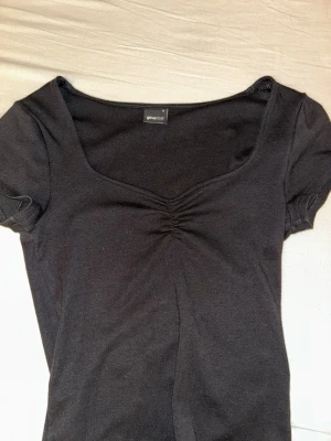 Svart topp med rynk från Gina Tricot - En svart topp från Gina Tricot med korta puffärmar och snygg rynkdetalj vid bysten. Toppen har en mjuk, stretchig känsla och en feminin v-ringad form. Perfekt att styla med jeans eller kjol för en trendig look.