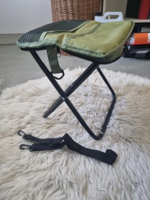 Hopfällbar grön campingpall - En smidig hopfällbar pall i grön textil med svart metallram. Sitsen är ca 30x25 cm och höjden är 30 cm. Perfekt att ta med på utflykter eller camping. Medföljer en svart axelrem med clips för enkel transport.