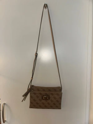 Guess väska - Snygg brun handväska med axelrem och dubbla dragkedjor. Väskan har ett elegant monogrammönster över hela ytan och flera fack inuti för kort och småsaker