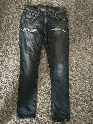 Nudie Mörkblå slitna jeans med hål - Säljer ett par mörkblå jeans med slitningar och stora hål vid knäna. Jeansen har en rak passform och klassisk femficksdesign. Perfekta för dig som gillar en edgy och avslappnad stil.