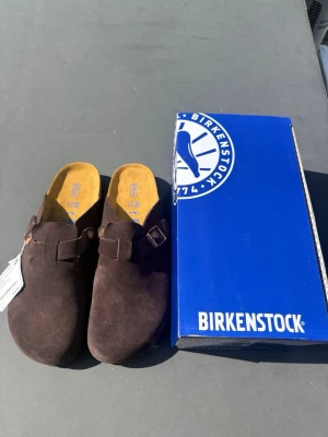 Bruna Birkenstock Boston - Säljer ett par Birkenstock tofflor i färgen mocha och är gjorda i mjuk mocka med justerbart spänne och klassisk korksula. Fotbädden är extra mjuk för skön komfort. Perfekta för dig som vill ha både stil och bekvämlighet. Hör av er vid eventuella frågor