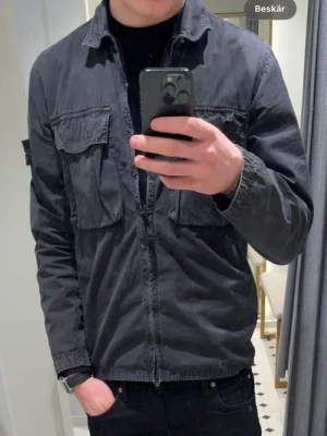 Stone Island overshirt  - Snygg svart overshirt från Stone Island med två stora bröstfickor och klassisk patch på vänster ärm. Overshirten har dragkedja framtill, krage och är tillverkad i ett slitstarkt bomullsmaterial. Perfekt för lager på lager och streetwear-stil.