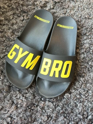Svarta slides GYMBRO Gymgrossisten - Säljer ett par svarta slides från Gymgrossisten med texten GYMBRO i stora gula bokstäver över remmen. Tillverkade i syntetmaterial med bekväm fotbädd och öppen tå. Perfekta för gymmet eller chill hemma.