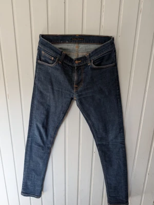 Mörkblå jeans från Nudie Jeans - Säljer ett par klassiska mörkblå jeans från Nudie Jeans med orangea kontrastsömmar och femficksmodell. Jeansen har rak passform och är tillverkade i slitstarkt denimtyg. Perfekta för dig som gillar stilrena och tidlösa jeans.