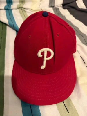 Röd Phillies keps från New Era - Snygg röd keps med vit broderad P-logga framtill, klassisk basebollstil från New Era. Kepsen har böjd skärm, ventilationshål och MLB-logga baktill. Insidan har svettband och märkning från 59FIFTY-serien.