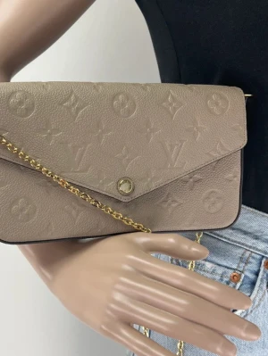 Beige Louis Vuitton handväska i läder - Säljer en beige handväska från Louis Vuitton i präglat läder med klassiskt monogrammönster. Väskan har guldfärgad kedjerem och kuvertformad design. Inuti finns avtagbara insatser för kort och mynt. Perfekt accessoar för dig som gillar lyxiga detaljer.