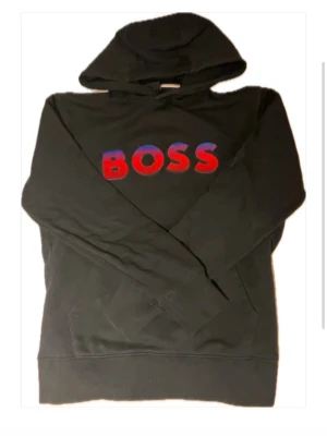 Svart hoodie från BOSS med röd-blå logga - Svart hoodie från BOSS med stor broderad logga i rött och blått på bröstet. Flekarna är bara på spegeln! Sitter bra och passar till massa olika outfits! Pris kan diskuteras! Tveka inte att höra av dig!