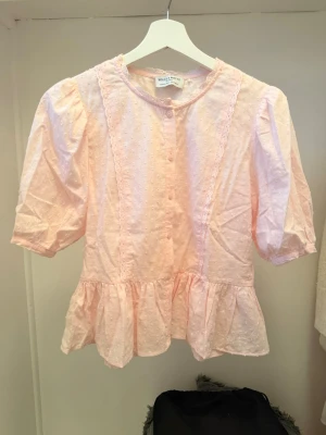 Ljusrosa blus med volang och puffärm - Supersöt ljusrosa blus med puffärmar, volang nedtill och knappar framtill. Blusen har diskret prickigt mönster i tyget och rund halsringning. Perfekt för dig som gillar romantisk och feminin stil.