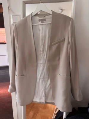Beige rak kavaj utan knappar - Stilren beige kavaj med rak passform och öppen front utan knappar. Kavajen har tre fickor fram och är helfodrad i ljust tyg. Perfekt för en clean och modern look. Materialet ser ut att vara en mjuk och slät textil.