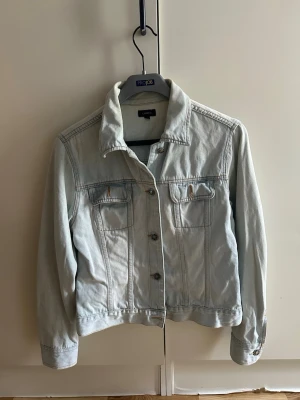 Ljusblå klassisk jeansjacka - Snygg ljusblå jeansjacka med raka sömmar och två bröstfickor med knapp. Jackan har en klassisk krage, knäppning framtill och justerbara knappar vid ärmslut och midja. Perfekt att slänga över en hoodie eller t-shirt för en avslappnad look.