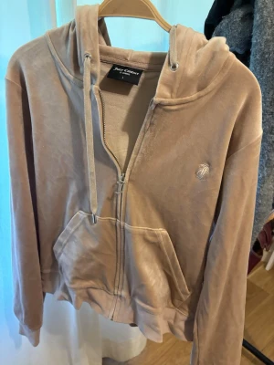 Beige velourhoodie Juicy Couture - Säljer en beige velourhoodie från Juicy Couture med dragkedja, huva och fickor framtill. Klassisk logga broderad på bröstet och mjukt material som känns lyxigt. Perfekt för en chill och trendig look.