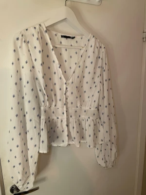 Vit blus med blått mönster från 157 - Söt vit blus från 157 med små blå blommönster, v-ringning och knappar framtill. Blusen har långärmade puffärmar och en volang nedtill som ger en luftig och romantisk känsla. Perfekt till jeans eller kjol för en fräsch look.