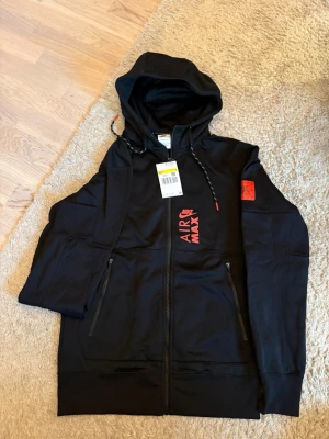 Svart Nike Air Max hoodie med dragkedja - Snygg svart hoodie från Nike Air Max med dragkedja framtill och två fickor med dragkedja. Röd Air Max-logga på bröstet och patch på ärmen. Justerbar huva med snören och mjukt material som känns skönt mot huden. Perfekt för en sportig och avslappnad stil.
