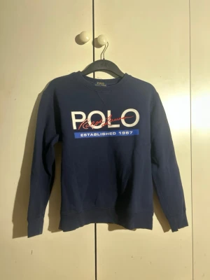 Blå sweatshirt från Polo Ralph Lauren - Mörkblå sweatshirt med rund hals från Polo Ralph Lauren. Stor logga framtill i vitt, rött och blått med texten 'POLO Ralph Lauren Established 1967'. Tröjan har långa ärmar och ribbade muddar vid ärmslut och nederkant.