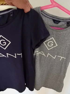 Två GANT t-shirts med logga - Säljer två snygga GANT t-shirts i storlek S. Ena är marinblå och den andra grå, båda med stor vit GANT-logga framtill. Klassisk rund hals och korta ärmar. Perfekta basplagg till garderoben och tillverkade i mjuk bomull. 150kr för båda