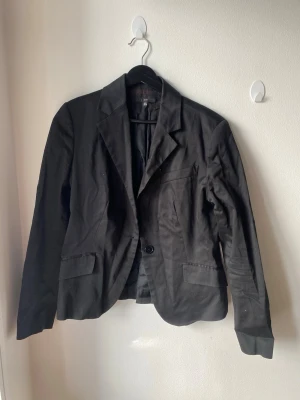Svart kavaj från ZARA - Stilren svart kavaj från ZARA med klassisk krage och enkelknäppning. Kavajen har två fickor med lock framtill och en bröstficka. Materialet är slätt och lätt, perfekt för en snygg och clean look.