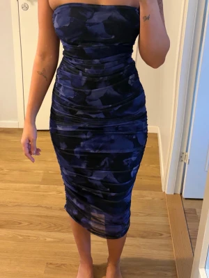 Marinblå axelbandslös långklänning - Säljer en marinblå axelbandslös långklänning med tight passform. Klänningen har ett abstrakt mönster i mörkblå toner och är figurnära med snygga draperingar längs sidorna. 