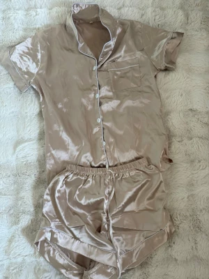 Beige satinpyjamas med kort ärm - Snygg beige pyjamas i glansigt satinmaterial med kortärmad skjorta och matchande shorts. Skjortan har vita knappar, bröstficka och kontrastsömmar. Perfekt för mysiga kvällar eller lata morgnar.