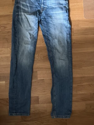 Blå raka jeans med slitningar - Säljer ett par klassiska blå jeans med raka ben och snygga slitningar framtill. Jeansen har normal midja och fem fickor. Perfekta för en avslappnad och trendig look. Materialet är denim i bomull. Längd 32. 28 i midjan 