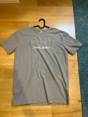 grå burberry t shirt - Snygg grå t-shirt från Burberry med vit och gul broderad text på bröstet och ett gult broderat emblem på ärmen. Klassisk passform och rund halsringning. Perfekt för dig som gillar stilrena plagg med exklusiva detaljer.
