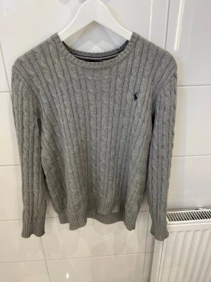 Grå kabelstickad tröja Polo Ralph Lauren - Kabelstickad grå tröja från Polo Ralph Lauren med rund halsringning och diskret broderad logga på bröstet. Tröjan har långa ärmar och ribbade muddar vid ärmslut och nederkant. Perfekt för lager på lager och klassisk stil. Den är i storleken 18-20 år vilket är ungefär XS till S